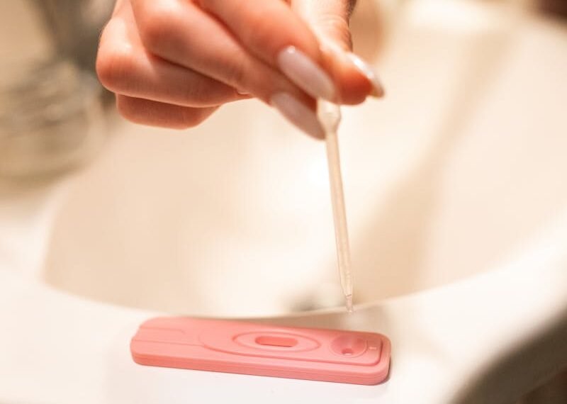 A hand using a pipette on a pregnancy test kit in a bathroom setting.